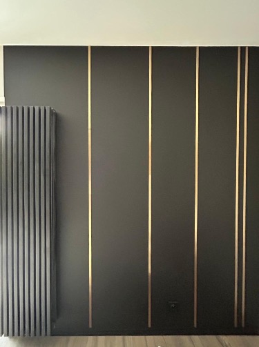 Ciemna ściana z dekoracyjnymi, złotymi listwami i grzejnikiem panelowym. Nowoczesny design wnętrza po remoncie.