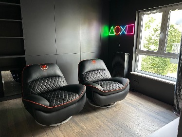 Dwa czarne, obrotowe fotele gamingowe z czerwonymi akcentami na tle szarego pokoju z neonowym logo PlayStation i oknem. Parkiet na podłodze.