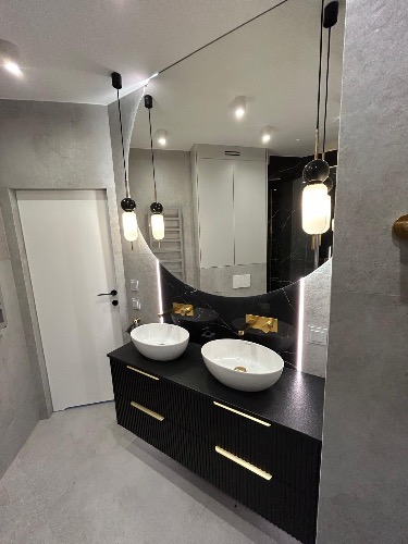 Nowoczesna łazienka z czarną szafką pod umywalki, okrągłym lustrem z podświetleniem LED i wiszącymi lampami. Minimalistyczny design, szare ściany.