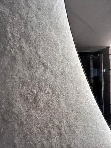 Szara ściana z wyraźną fakturą kamienia w łazience, odbicie kabiny prysznicowej w lustrze. Minimalistyczny design, nowoczesne wykończenie.