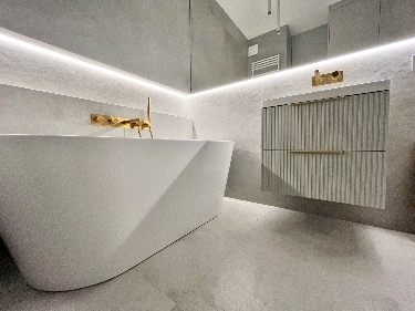 Minimalistyczna łazienka: biała wanna wolnostojąca, złota armatura, szafka z lamelami i podświetleniem LED. Szare ściany z efektem betonu.
