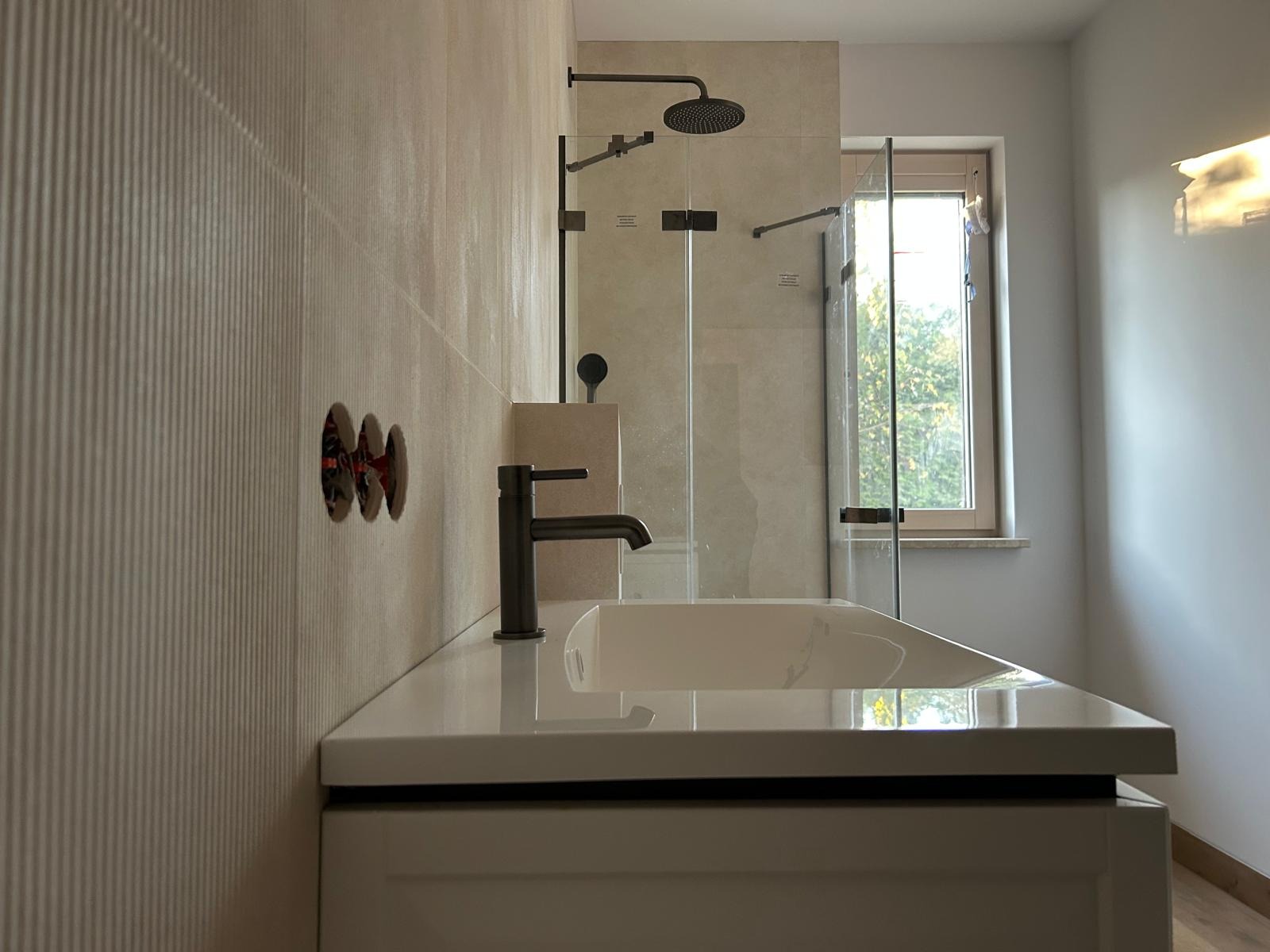 Nowoczesna łazienka z beżowymi płytkami, czarną armaturą i kabiną prysznicową. Widoczne otwory na baterie w ścianie i fragment okna z widokiem na zieleń.