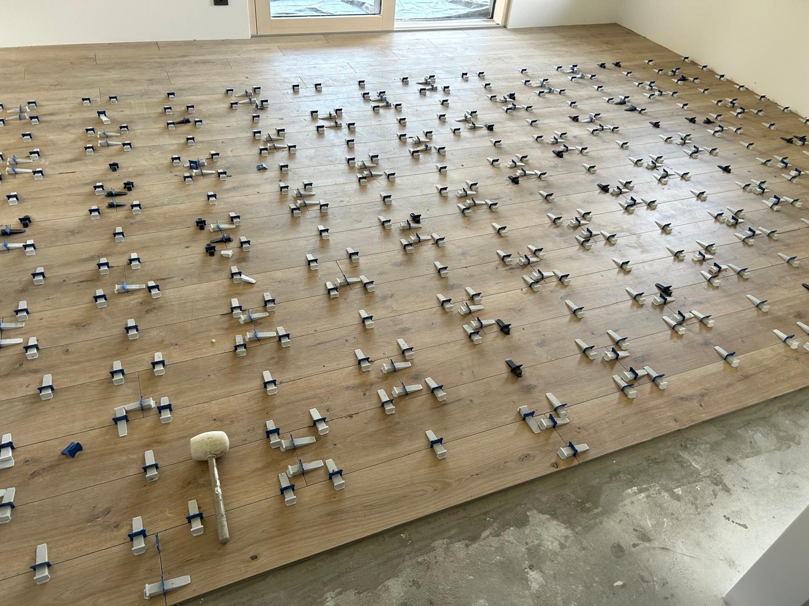 Układanie paneli podłogowych z użyciem klipsów poziomujących i gumowego młotka. Widok z góry na jasne panele imitujące drewno w trakcie montażu.