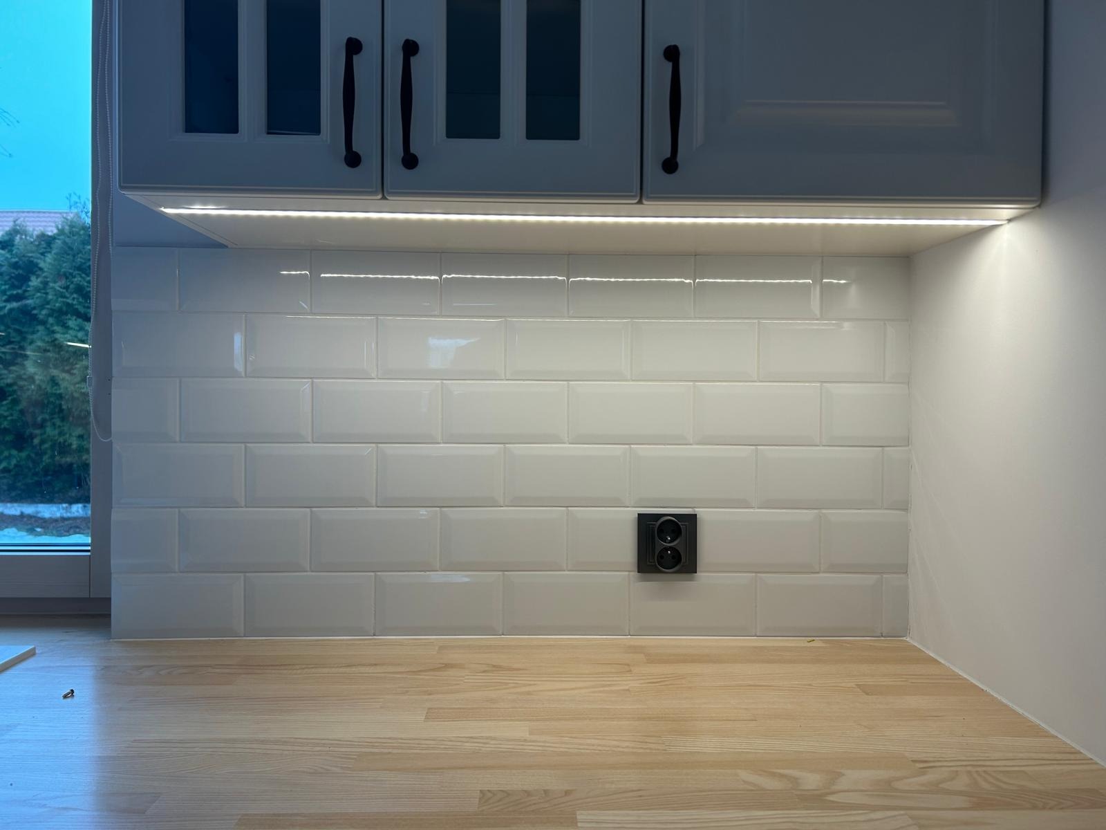 Białe, błyszczące płytki typu metro na ścianie nad drewnianym blatem kuchennym, oświetlone listwą LED pod szafkami. Widoczne gniazdko elektryczne.