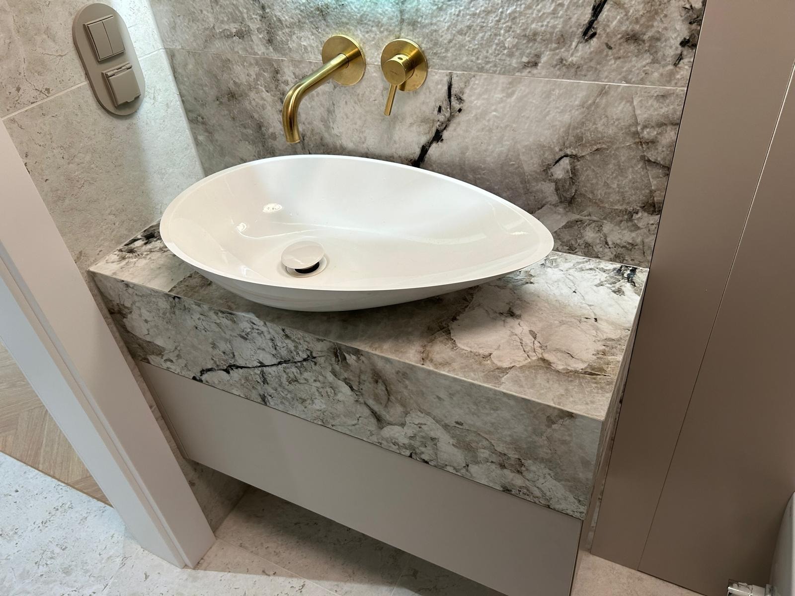 Biała, owalna umywalka nablatowa z baterią w kolorze złotym, osadzona na blacie z marmuru w łazience. Widoczne płytki imitujące marmur na ścianie.
