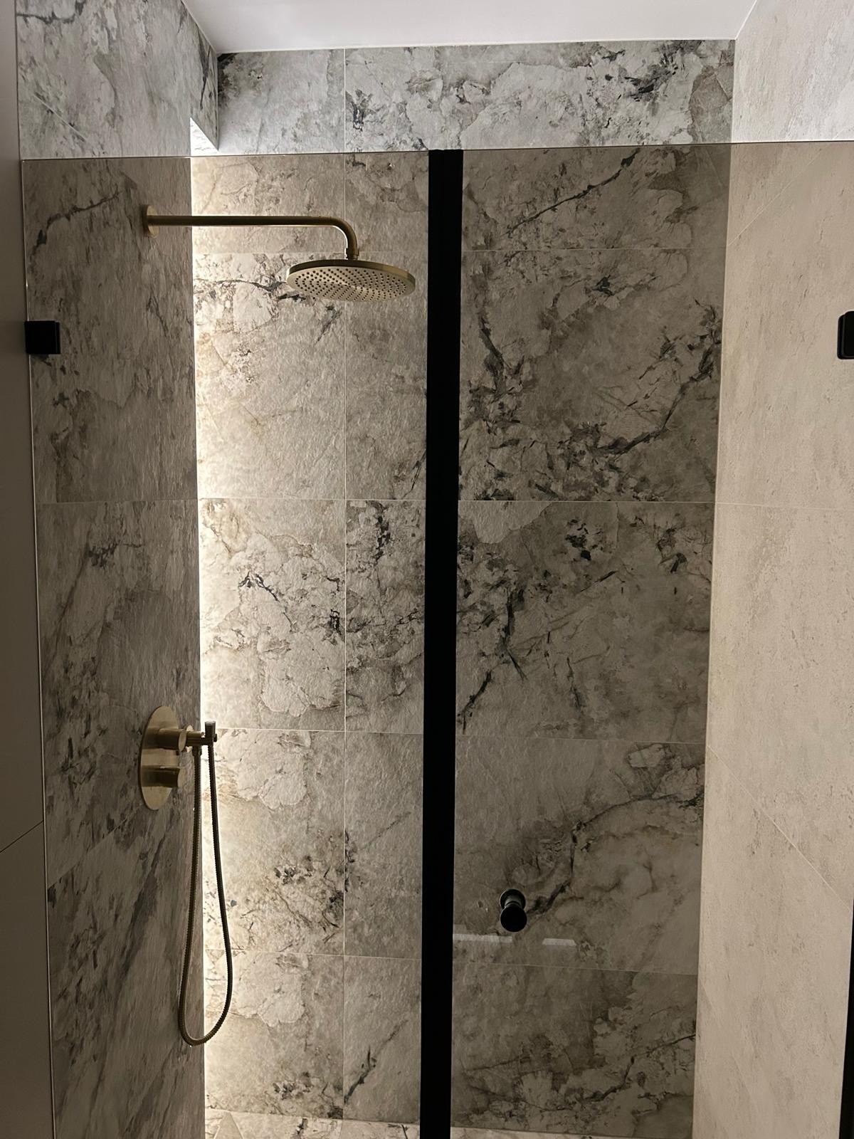 Nowoczesna kabina prysznicowa z marmurowymi płytkami i złotą armaturą. Czarny, pionowy akcent oddziela strefy. Minimalistyczny design, eleganckie wykończenie.