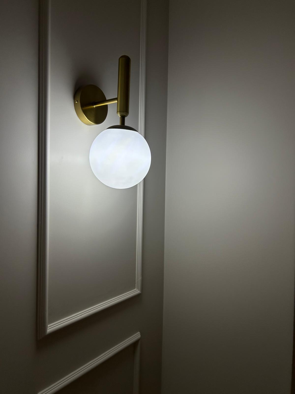 Elegancki kinkiet z kulistym, mlecznym kloszem i złotym wykończeniem, zamontowany na ścianie z dekoracyjnymi listwami. Subtelne oświetlenie wnętrza.