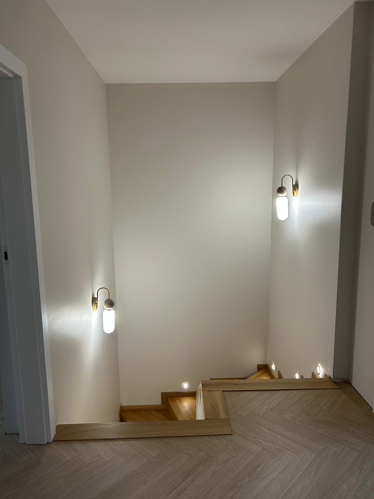 Nowoczesne schody z drewnianymi stopniami i oświetleniem punktowym w ścianie, wykończone w jasnych barwach, z parkietem ułożonym w jodełkę.