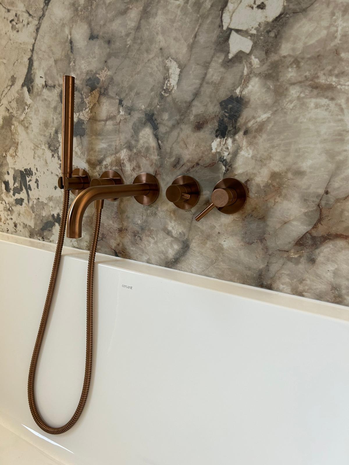 Miedziana bateria wannowa na tle marmurowej ściany, nowoczesny design, widoczny fragment białej wanny. Eleganckie wykończenie łazienki.