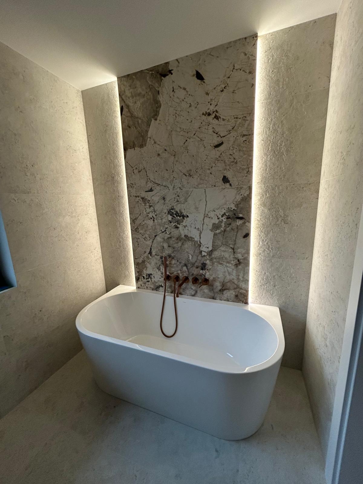 Nowoczesna łazienka z wolnostojącą wanną, marmurową ścianą akcentową i subtelnym oświetleniem LED. Armatura w kolorze miedzi dodaje elegancji.