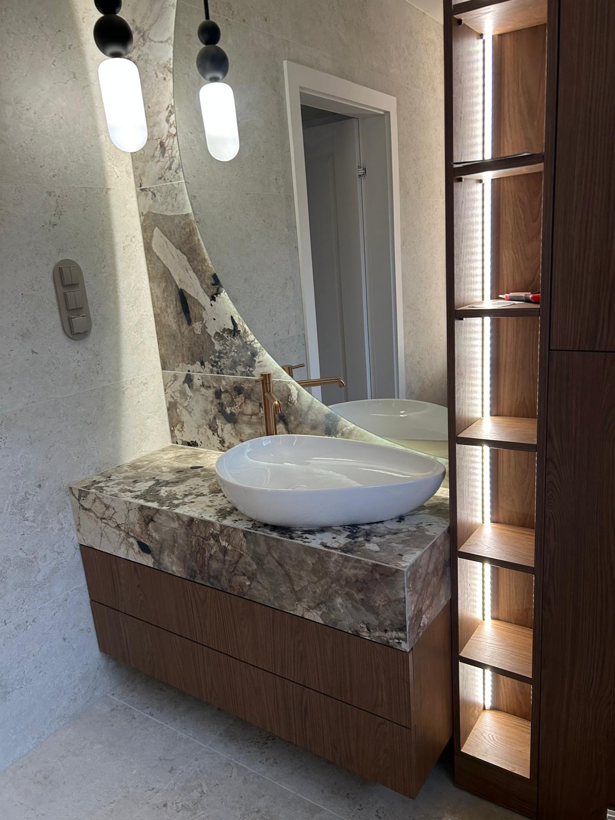 Nowoczesna łazienka z umywalką nablatową na blacie z marmuru, podświetlane półki i okrągłe lustro z dekoracyjnym wzorem. Armatura w kolorze miedzi.