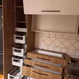 Montaż szuflad cargo w szafce kuchennej z frontami w kolorze drewna i białymi korpusami, widoczne elementy instalacji elektrycznej na ścianie.