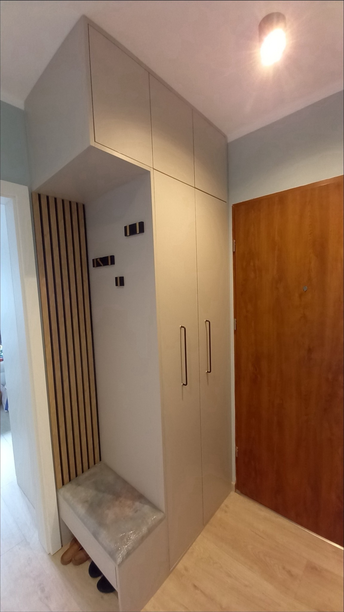 Beżowa zabudowa przedpokoju z szafą, siedziskiem i drewnianymi lamelami, uzupełniona o minimalistyczne wieszaki i brązowe drzwi wejściowe.