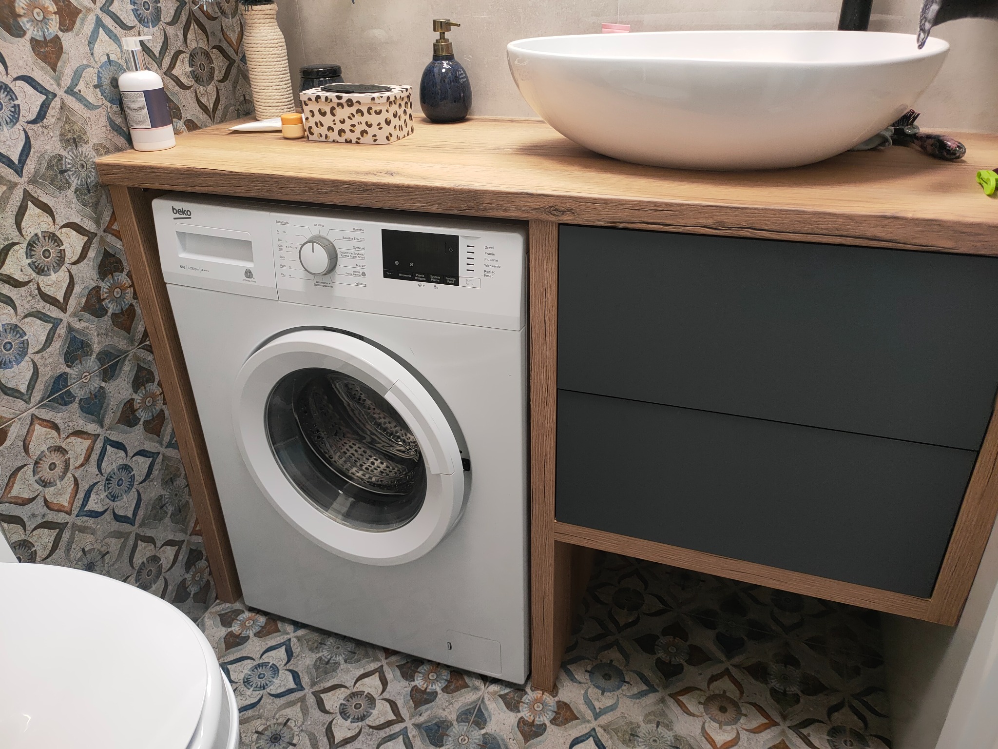 Aranżacja łazienki z pralką Beko w zabudowie pod blatem umywalki nablatowej i szafką z szufladami, całość w tonacji drewna i grafitu, na tle wzorzystych płytek podłogowych.