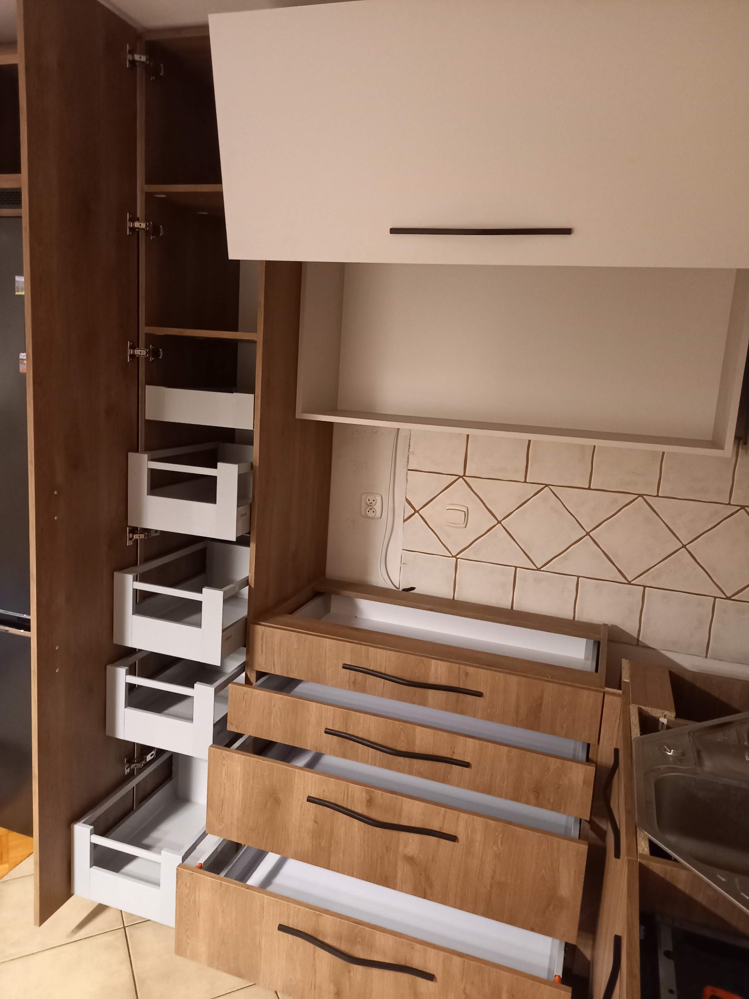 Montaż szuflad cargo w szafce kuchennej z frontami w kolorze drewna i białymi korpusami, widoczne elementy instalacji elektrycznej na ścianie.