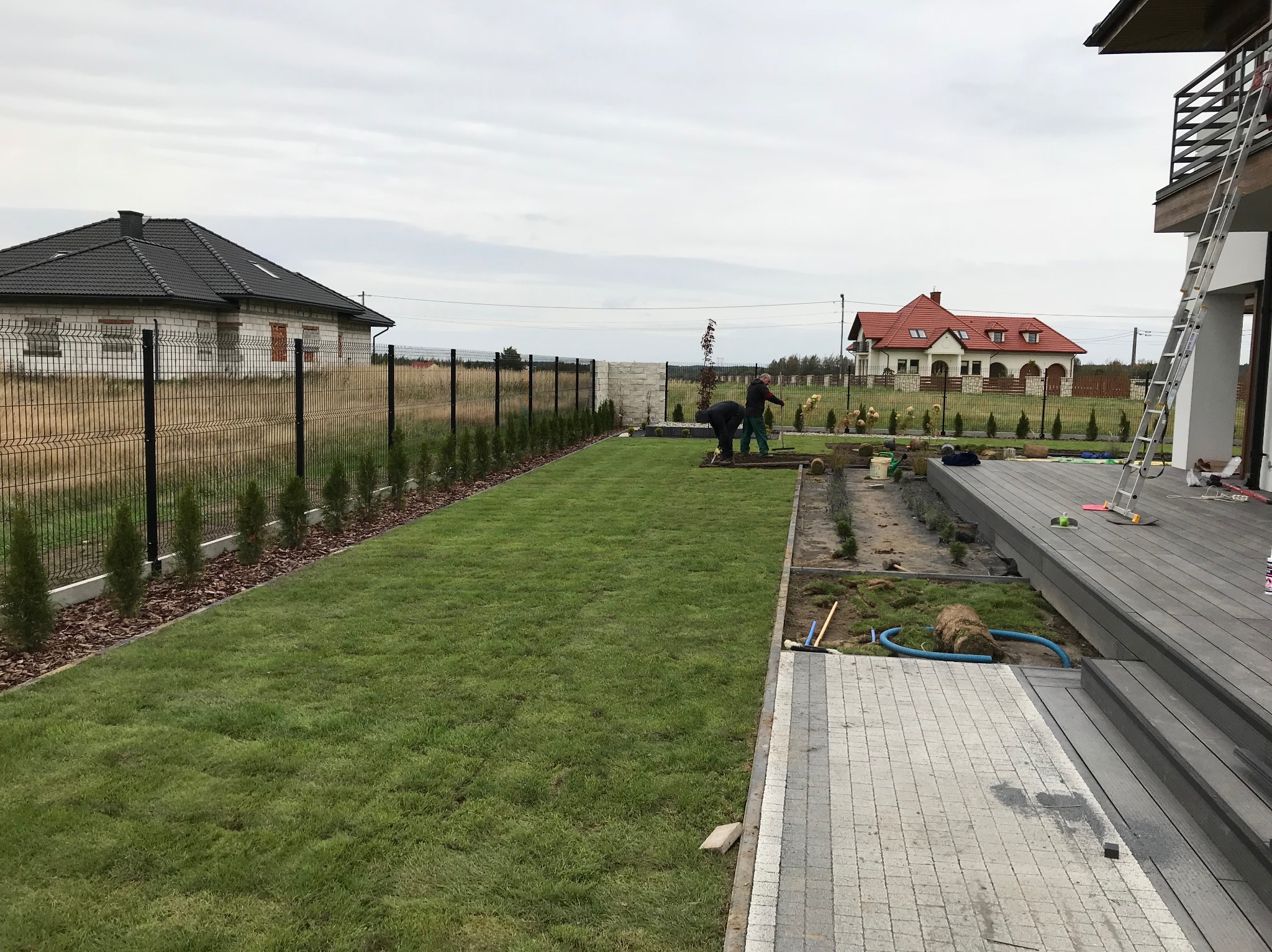 Widok na ogród w trakcie zakładania: trawnik z rolki, przygotowane rabaty, pracownicy w trakcie sadzenia roślin, w tle domy i ogrodzenie.