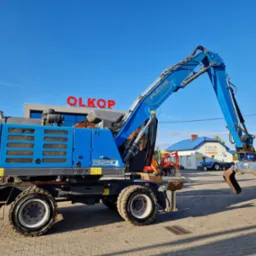 Niebieska koparka Fuchs 320 z chwytakiem hydraulicznym na tle błękitnego nieba i budynków stacji paliw OLKOP.