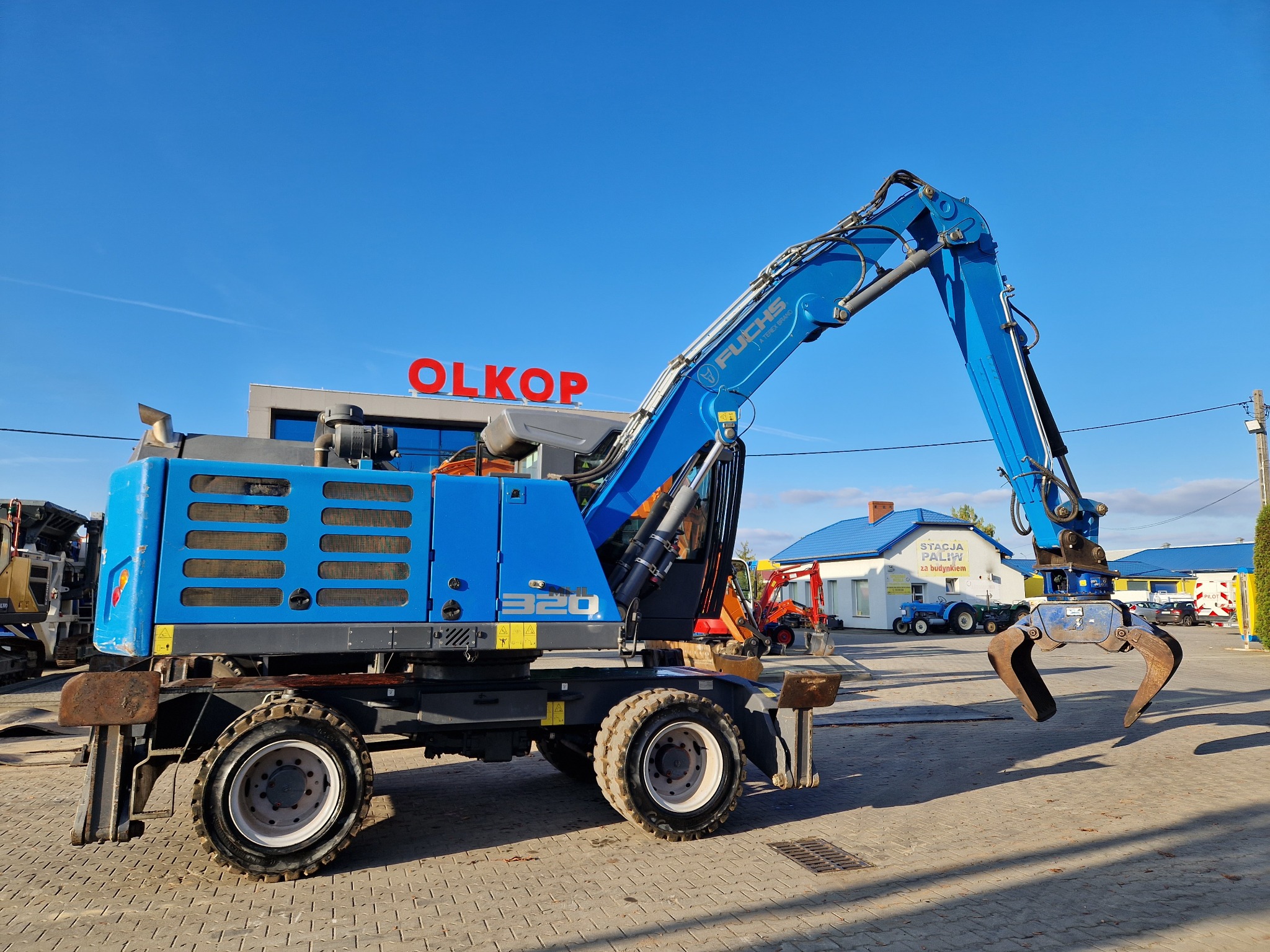 Niebieska koparka Fuchs 320 z chwytakiem hydraulicznym na tle błękitnego nieba i budynków stacji paliw OLKOP.