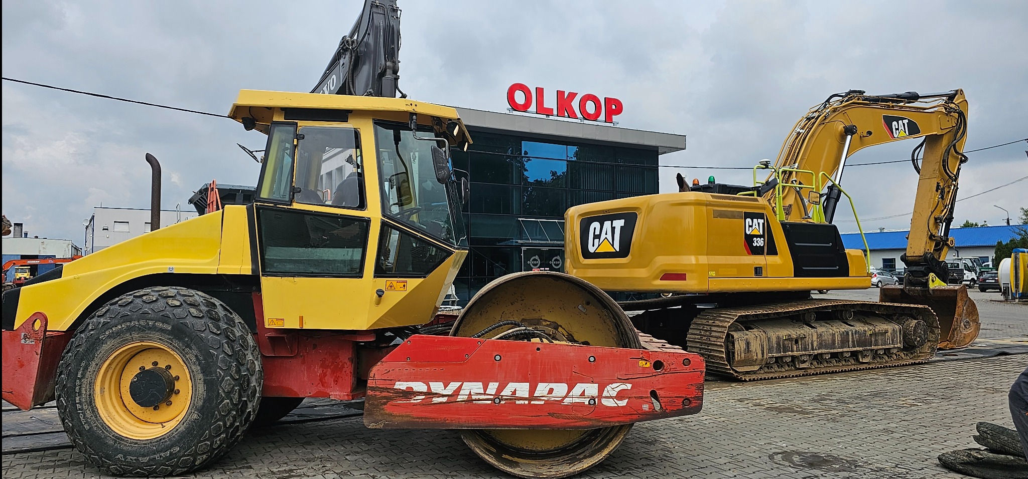 Żółty walec drogowy Dynapac i koparka CAT 336 na placu firmy OLKOP, widoczne ślady użytkowania na sprzęcie.