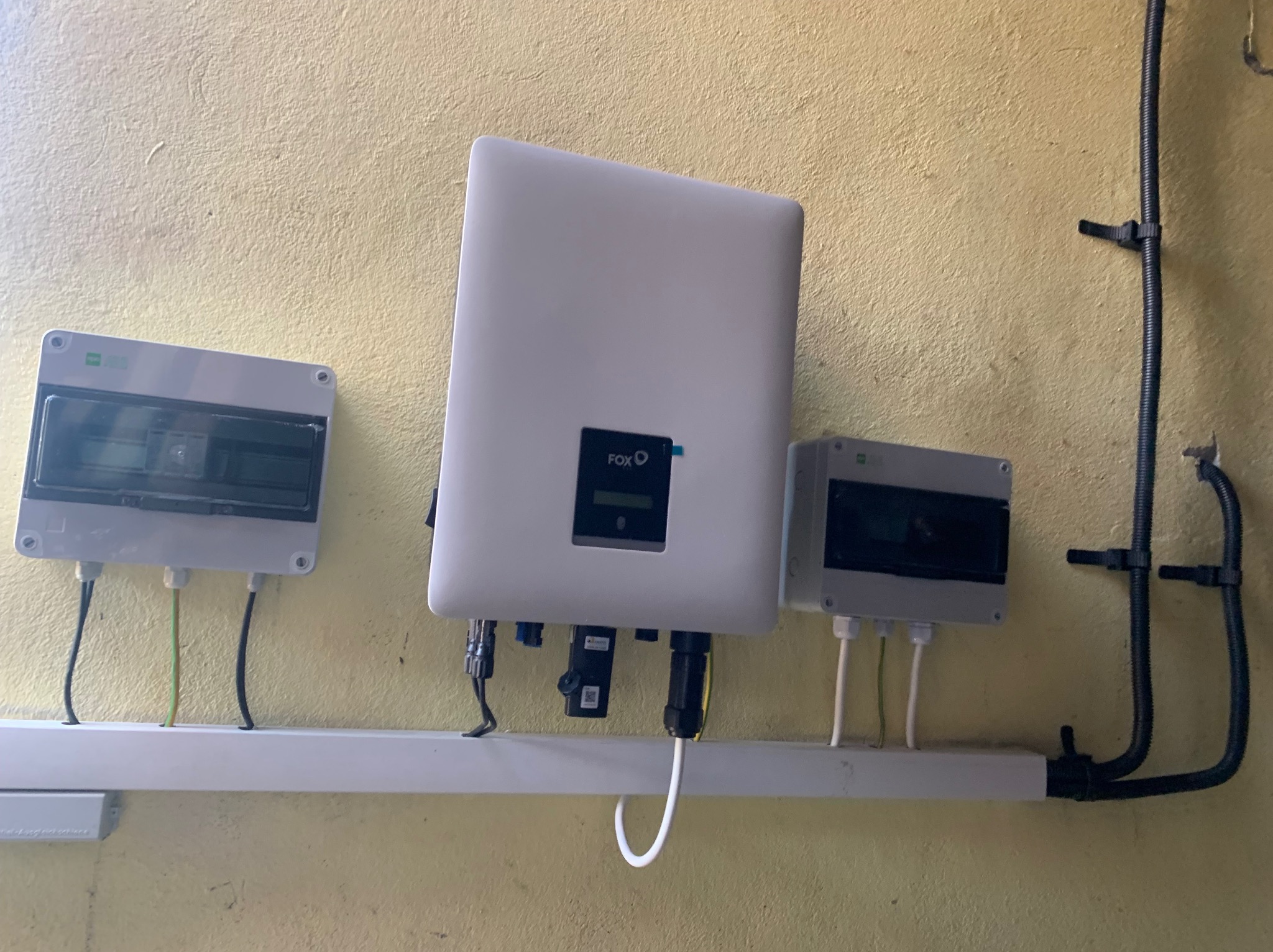 Biały inwerter marki Fox oraz dwie szare skrzynki rozdzielcze zamontowane na ścianie o szorstkiej fakturze, widoczne kable i przewody elektryczne.