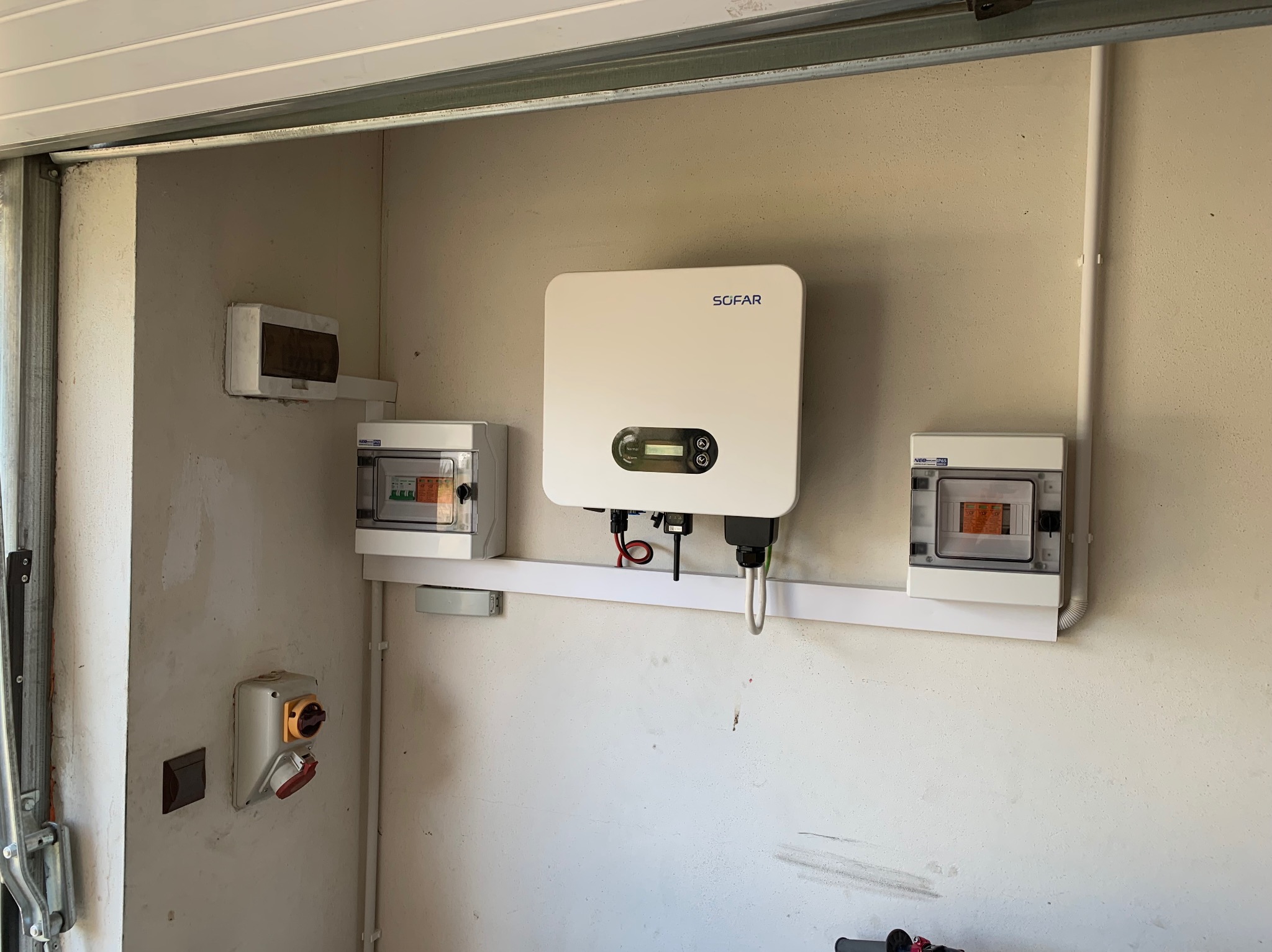 Zamontowany inwerter SOFAR i skrzynki zabezpieczeń na ścianie w garażu, widoczne przewody i elementy instalacji elektrycznej.