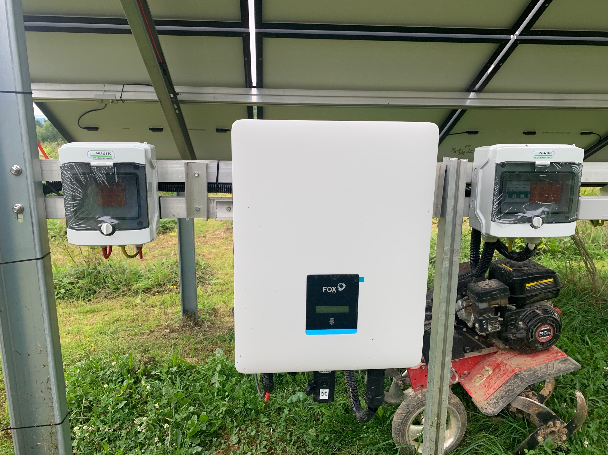 Instalacja z inwerterem FOX ESS i zabezpieczeniami Noark, widoczna glebogryzarka spalinowa na trawiastym terenie.