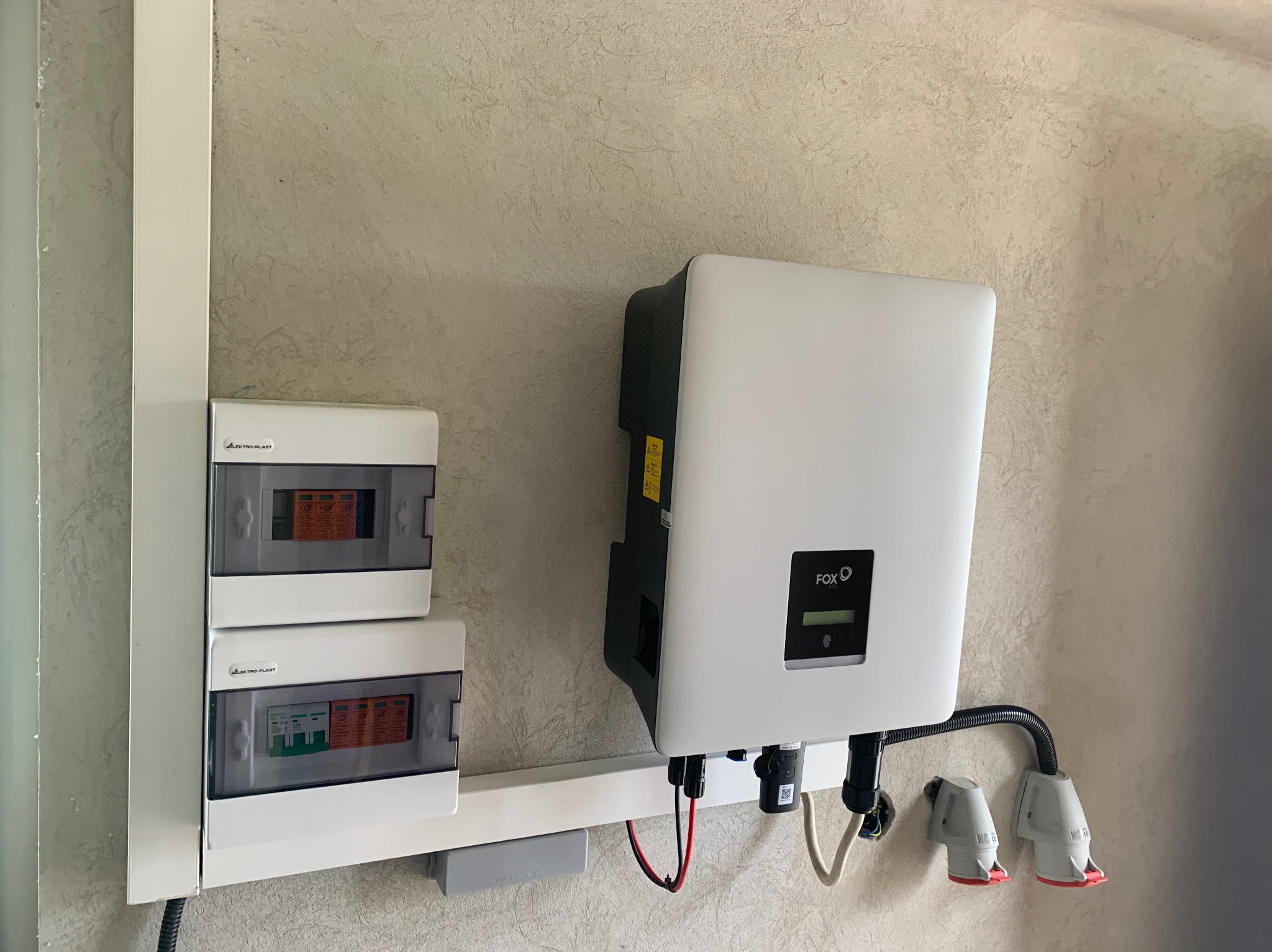 Biała skrzynka inwertera FOX ESS zamontowana na ścianie z dwoma skrzynkami bezpieczników Elektro-Plast, widoczne przewody i złącza.