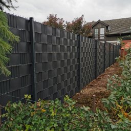 Baza-zaune - Długi, grafitowy system ogrodzeniowy z paneli kompozytowych o wzorze szachownicy, oddzielający posesję od sąsiadów, z widoczną zielenią w tle i pnączami bluszczu u podstawy.