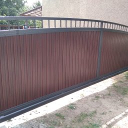 Baza-zaune - Przesuwna brama wjazdowa z brązowymi panelami pionowymi i szarą metalową ramą, widoczna z boku na tle trawnika i budynku.