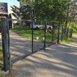 Baza-zaune - Czarna, dwuskrzydłowa brama z siatki z widocznym mechanizmem zamykającym, zamontowana na betonowej podmurówce, z fragmentem siatki ogrodzeniowej w tle, obok zaparkowany bus z pracownikiem w środku.