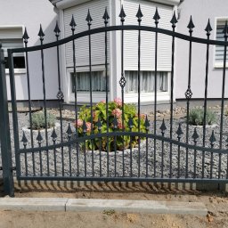 Baza-zaune - Eleganckie, czarne ogrodzenie z metalu z dekoracyjnymi grotami, widoczne na tle jasnego domu z żaluzjami w oknach, otoczone ozdobnym żwirem i roślinnością.