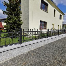 Baza-zaune - Eleganckie, czarne ogrodzenie kute z ozdobnymi elementami, zamontowane na niskim murku z szarej kostki, na tle domu z kremową elewacją i zielenią ogrodu.