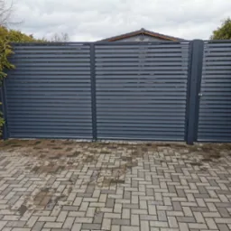 Nowoczesna brama wjazdowa z poziomych, szarych paneli, widoczna z zewnątrz na tle zielonych tui i kostki brukowej. Po lewej stronie zielona konewka.