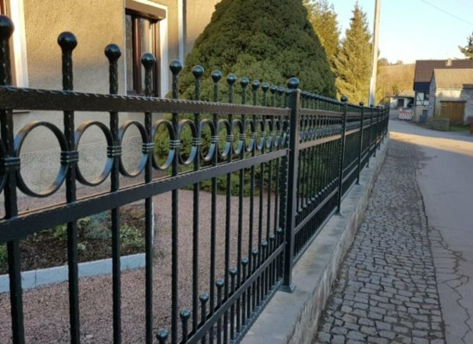 Czarne, metalowe ogrodzenie z okrągłymi dekoracjami na tle domu i zieleni.