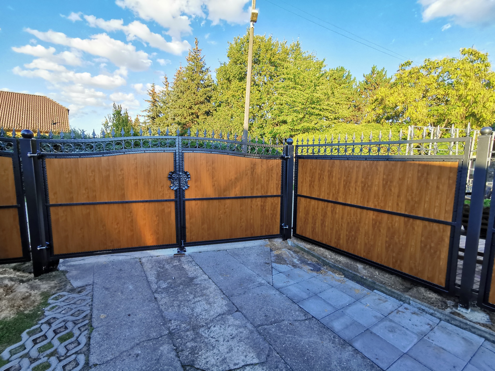 Otwarta brama wjazdowa dwuskrzydłowa z drewnopodobnym wypełnieniem i kutymi zdobieniami, widok na posesję z kostką brukową i drzewami w tle.
