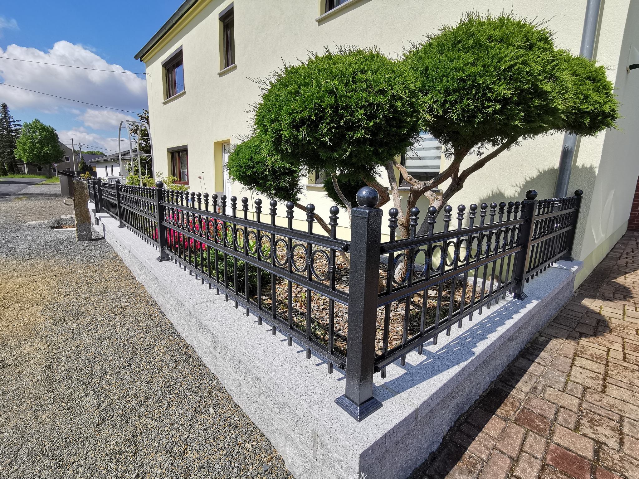Czarny, metalowy płot z ozdobnymi okręgami, osadzony na niskim, kamiennym murku, otaczający posesję z zielonymi krzewami i żółtym domem w tle.