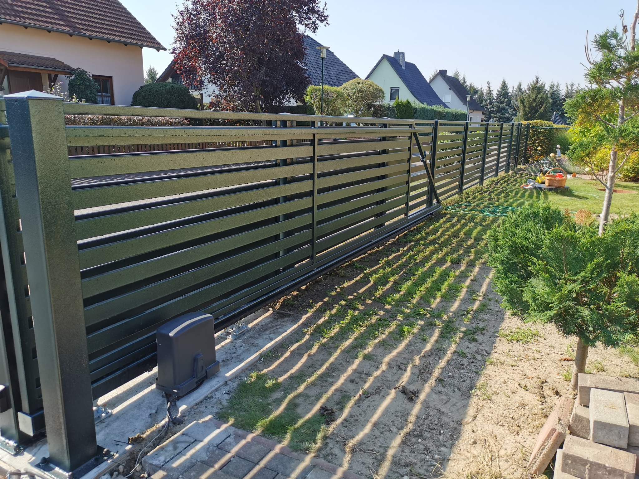 Nowoczesna przesuwna brama wjazdowa z poziomych paneli metalowych, wyposażona w napęd elektryczny, widoczna z zewnątrz posesji na tle domów jednorodzinnych.