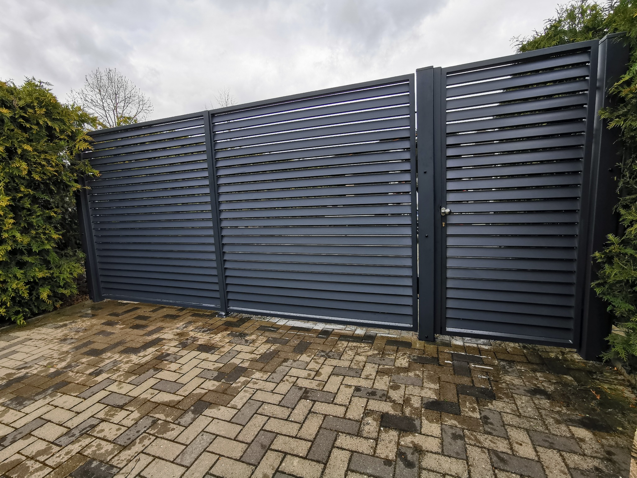 Nowoczesna, pozioma brama wjazdowa z szarego metalu, z widocznymi poziomymi lamelami, na tle kostki brukowej ułożonej w jodełkę i zielonych krzewów.
