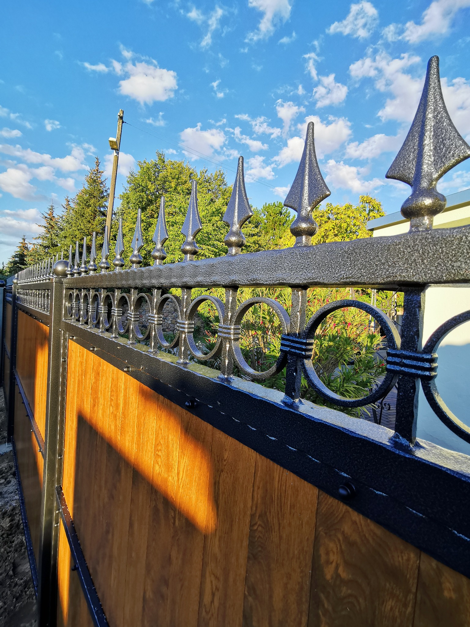 Fragment metalowej bramy wjazdowej z drewnianym wypełnieniem, zakończonej ozdobnymi grotami, oświetlonej popołudniowym słońcem, na tle błękitnego nieba z chmurami.