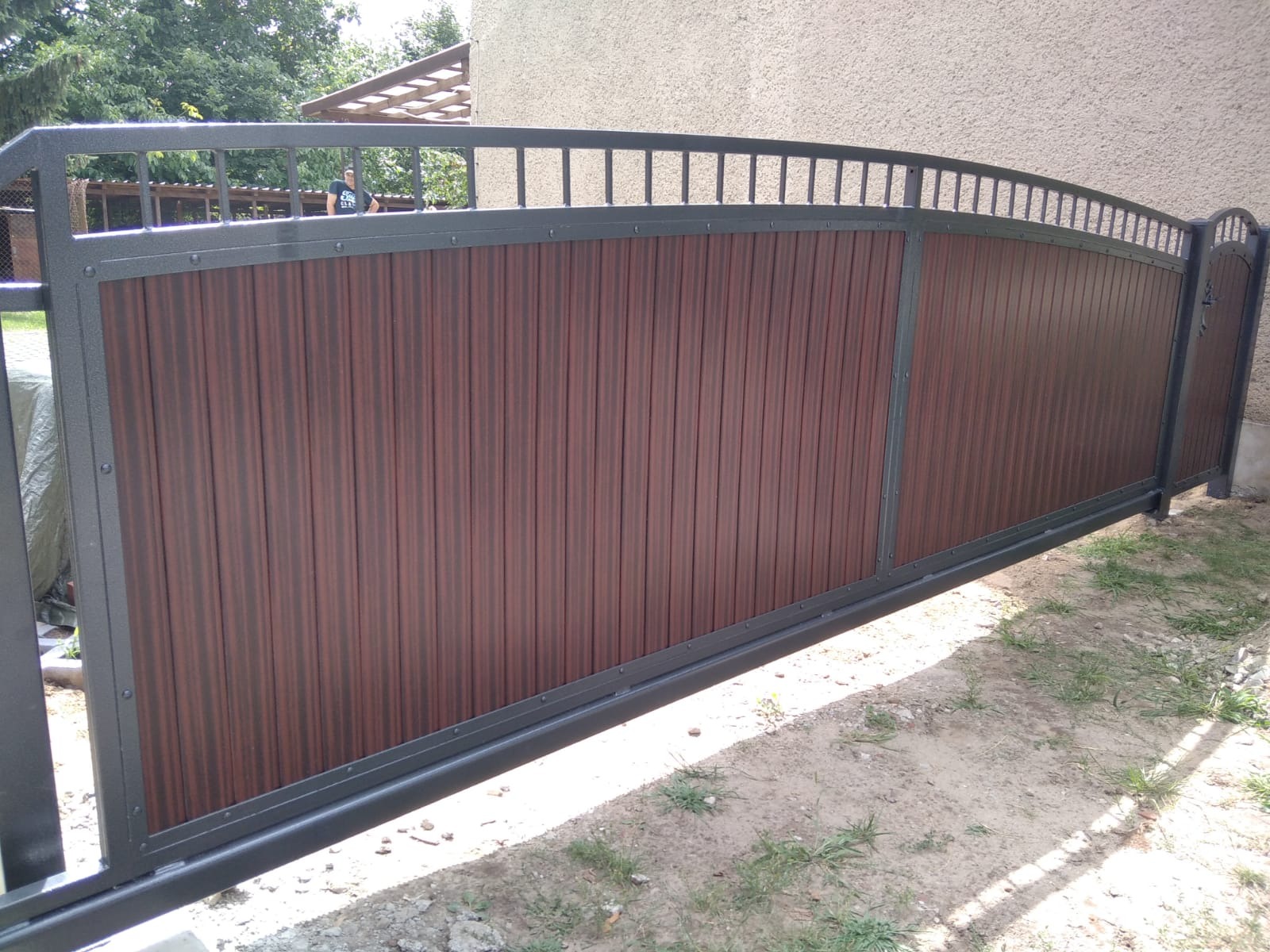 Przesuwna brama wjazdowa z brązowymi panelami pionowymi i szarą metalową ramą, widoczna z boku na tle trawnika i budynku.