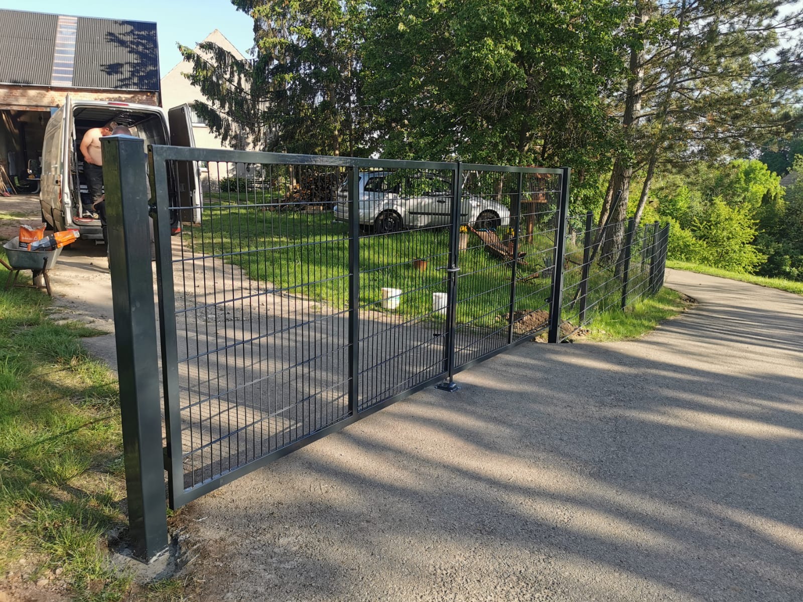 Czarna, dwuskrzydłowa brama z siatki z widocznym mechanizmem zamykającym, zamontowana na betonowej podmurówce, z fragmentem siatki ogrodzeniowej w tle, obok zaparkowany bus z pracownikiem w środku.