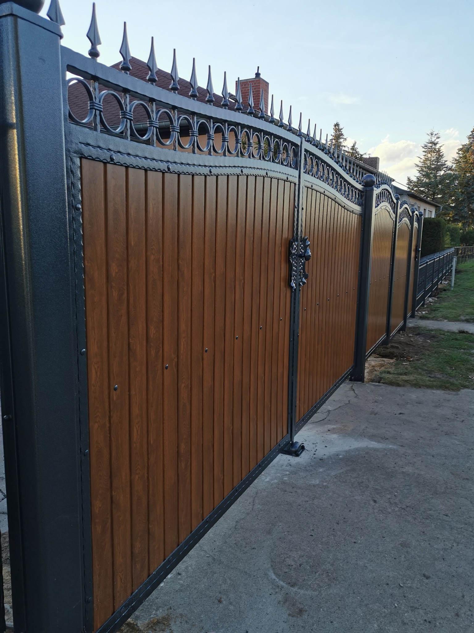 Nowoczesna brama wjazdowa z drewnianymi panelami i metalową konstrukcją z ozdobnymi elementami, widok z boku na betonowy podjazd.