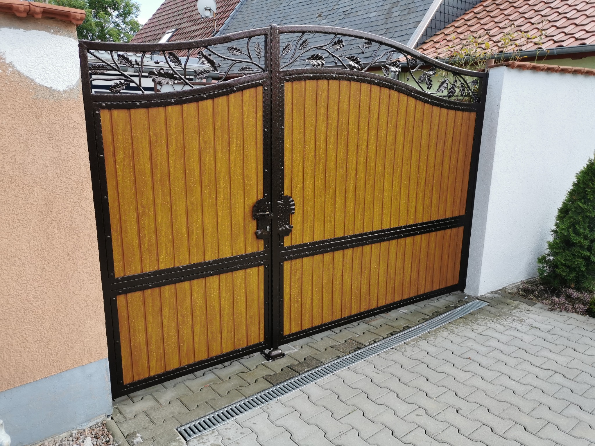 Dwuskrzydłowa brama wjazdowa z drewnianymi panelami w kolorze miodowym i ozdobnym metalowym wykończeniem z motywem liści, zamontowana na posesji z kostką brukową i odpływem liniowym.