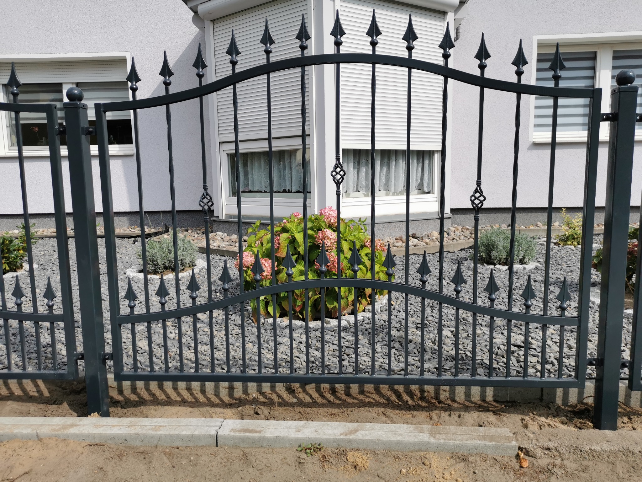 Eleganckie, czarne ogrodzenie z metalu z dekoracyjnymi grotami, widoczne na tle jasnego domu z żaluzjami w oknach, otoczone ozdobnym żwirem i roślinnością.