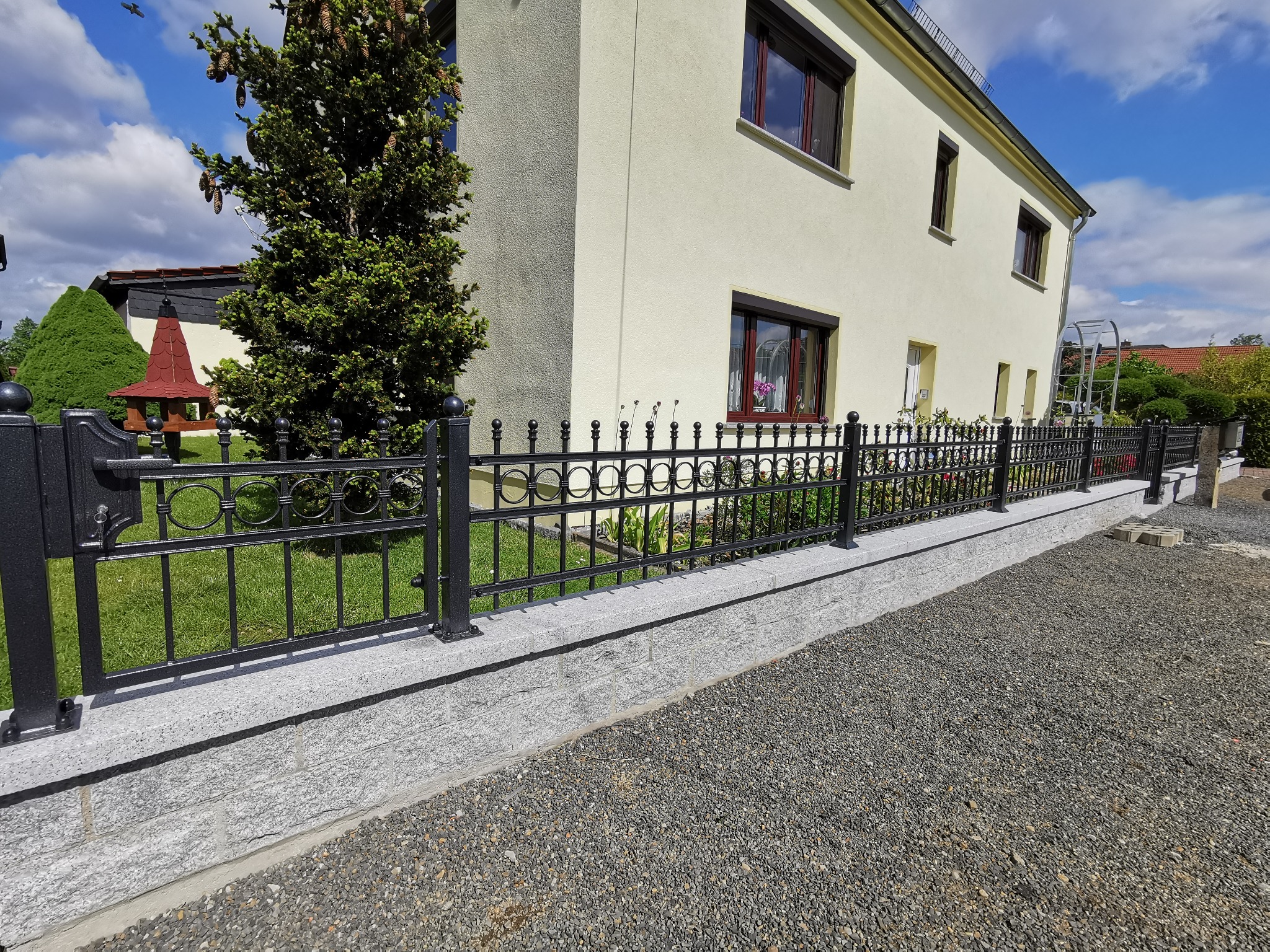 Eleganckie, czarne ogrodzenie kute z ozdobnymi elementami, zamontowane na niskim murku z szarej kostki, na tle domu z kremową elewacją i zielenią ogrodu.