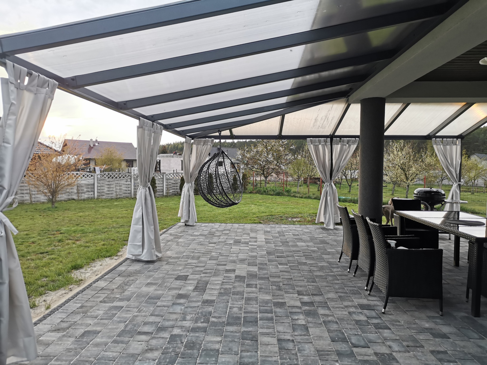 Zadaszony taras z szarą kostką brukową, wiszącym fotelem-kokonem, zasłonami i meblami ogrodowymi, widok na ogród i dom w tle.