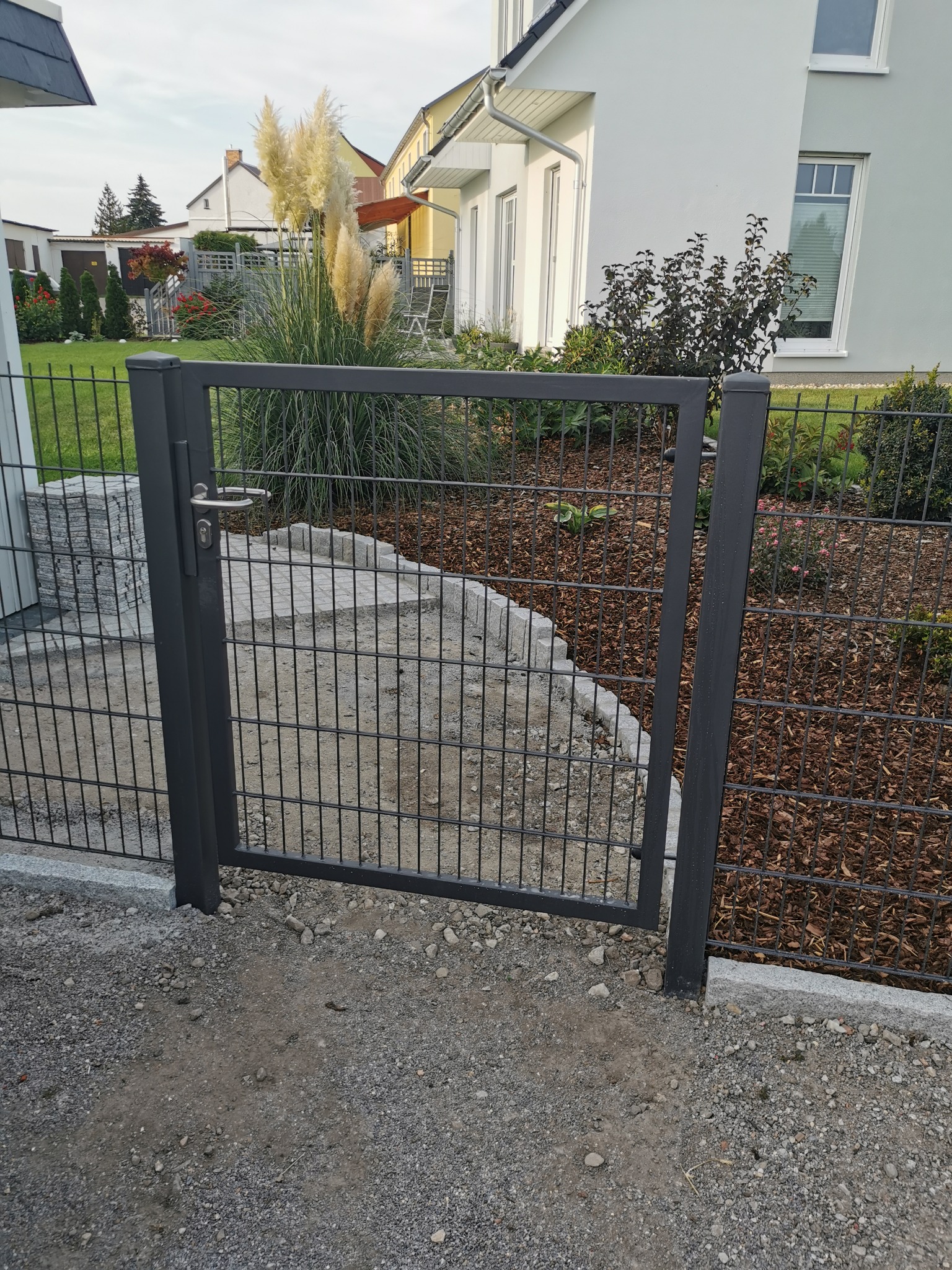 Nowoczesna furtka z paneli ogrodzeniowych w kolorze grafitowym, z widoczną klamką i zamkiem, prowadząca do ogrodu z korą i ozdobną trawą pampasową, na tle jasnego domu.