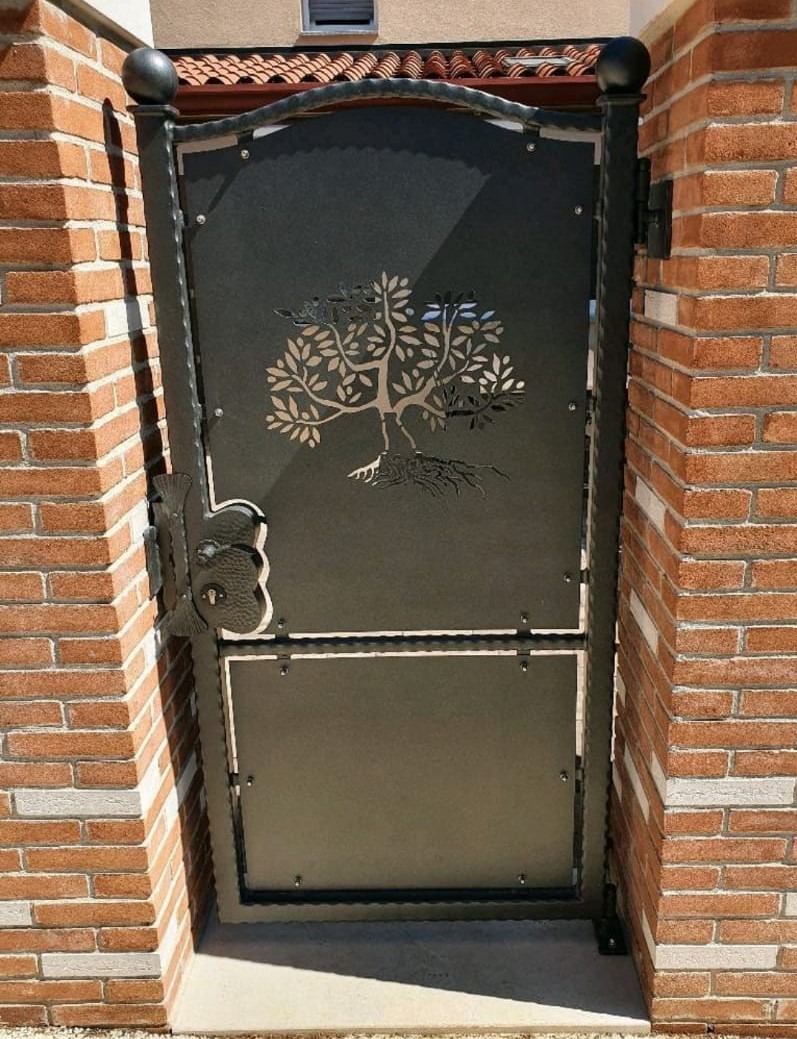 Kuta, metalowa furtka z wyciętym motywem drzewa, zamontowana w murze z czerwonej cegły.