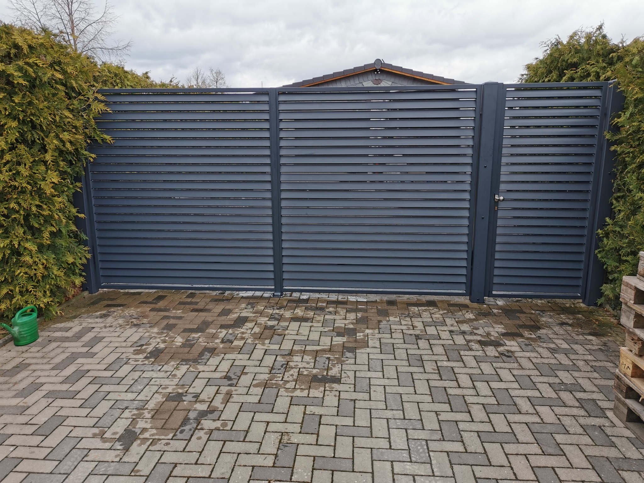 Nowoczesna brama wjazdowa z poziomych, szarych paneli, widoczna z zewnątrz na tle zielonych tui i kostki brukowej. Po lewej stronie zielona konewka.