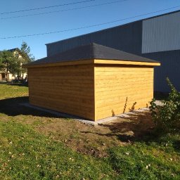 Piotr łozowski alt- drew - Niewielki, świeżo zaimpregnowany, drewniany budynek z czarnym, spadzistym dachem, ustawiony na betonowej podstawie na trawniku, z budynkiem gospodarczym w tle.